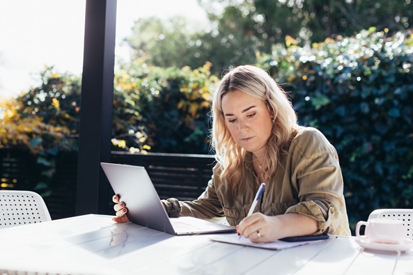 A young woman with blond here is sitting outside at a table writing notes with her laptop open. We see trees and the sky in the background.