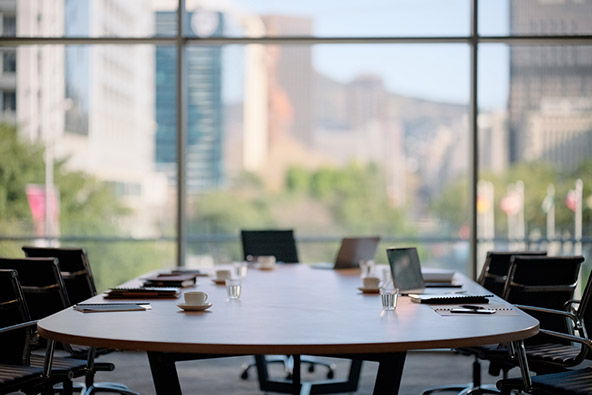A long board table with papers, laptops, tea cups and glasses of water on its surface. Empty chairs surround the table. In the background we see trees and high rise buildings