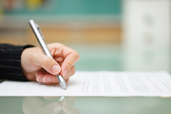 A left hand holding a silver pen and signing a paper document