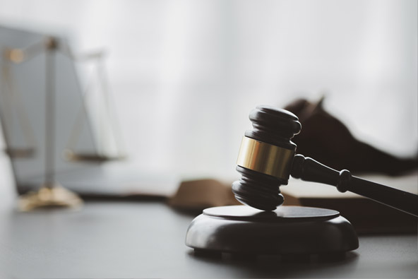 A wooden judge's gavel and block sit on a desk. In the background, out of focus, is a gold legal scale and a hand holding a pen
